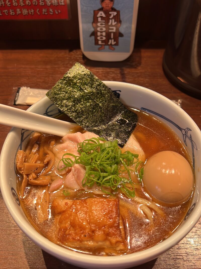 麵屋武蔵の武蔵ら～麺あっさり、の画像。醤油色のスープにうっすら浮いた脂が光る。
トッピングは上から時計回りに、海苔、煮卵、角煮、メンマ、中心にはピンク色の薄切りチャーシューと彩の青ネギ